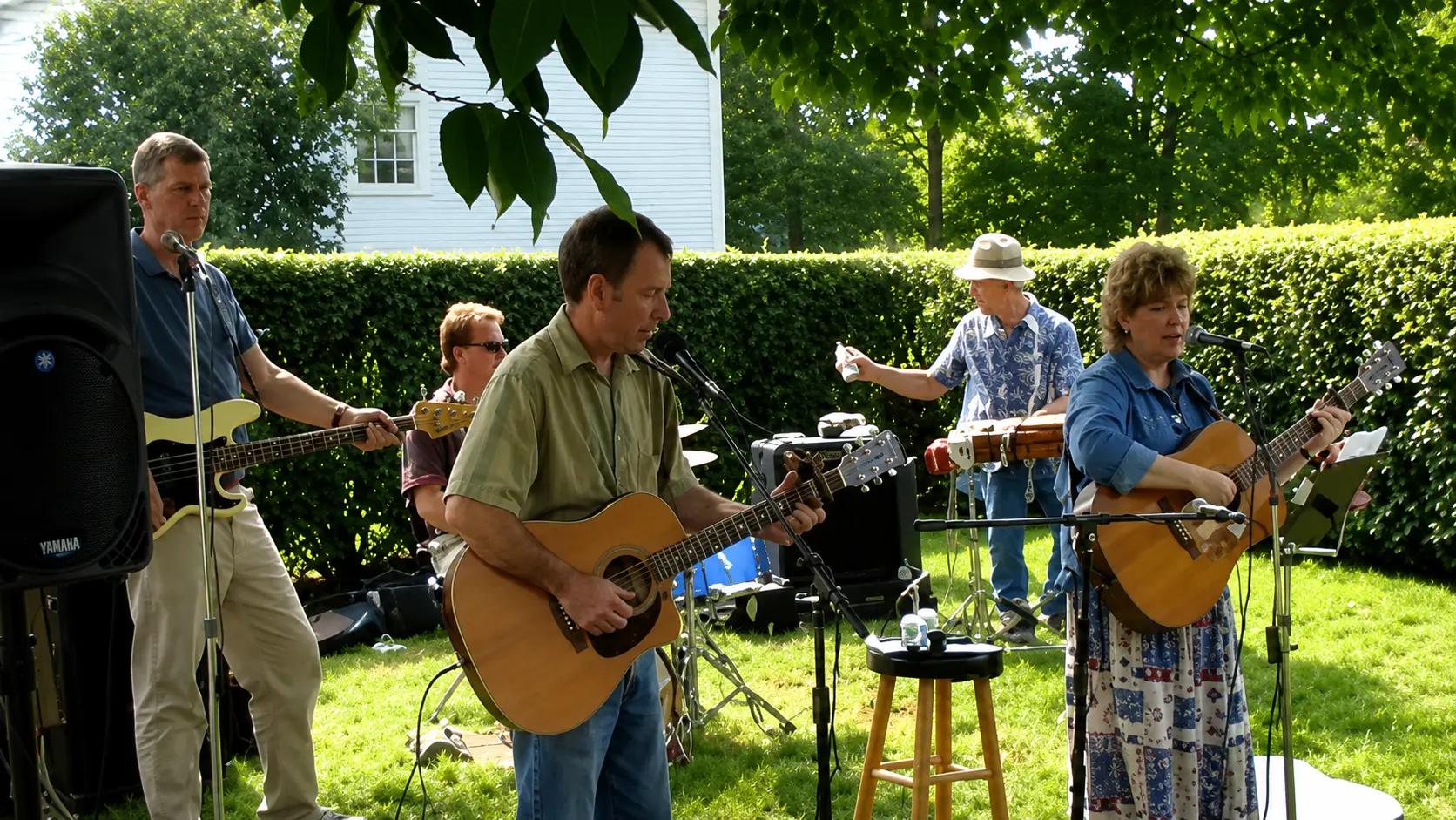 Three musicians playing guitars and percussion in a sunny backyard garden
